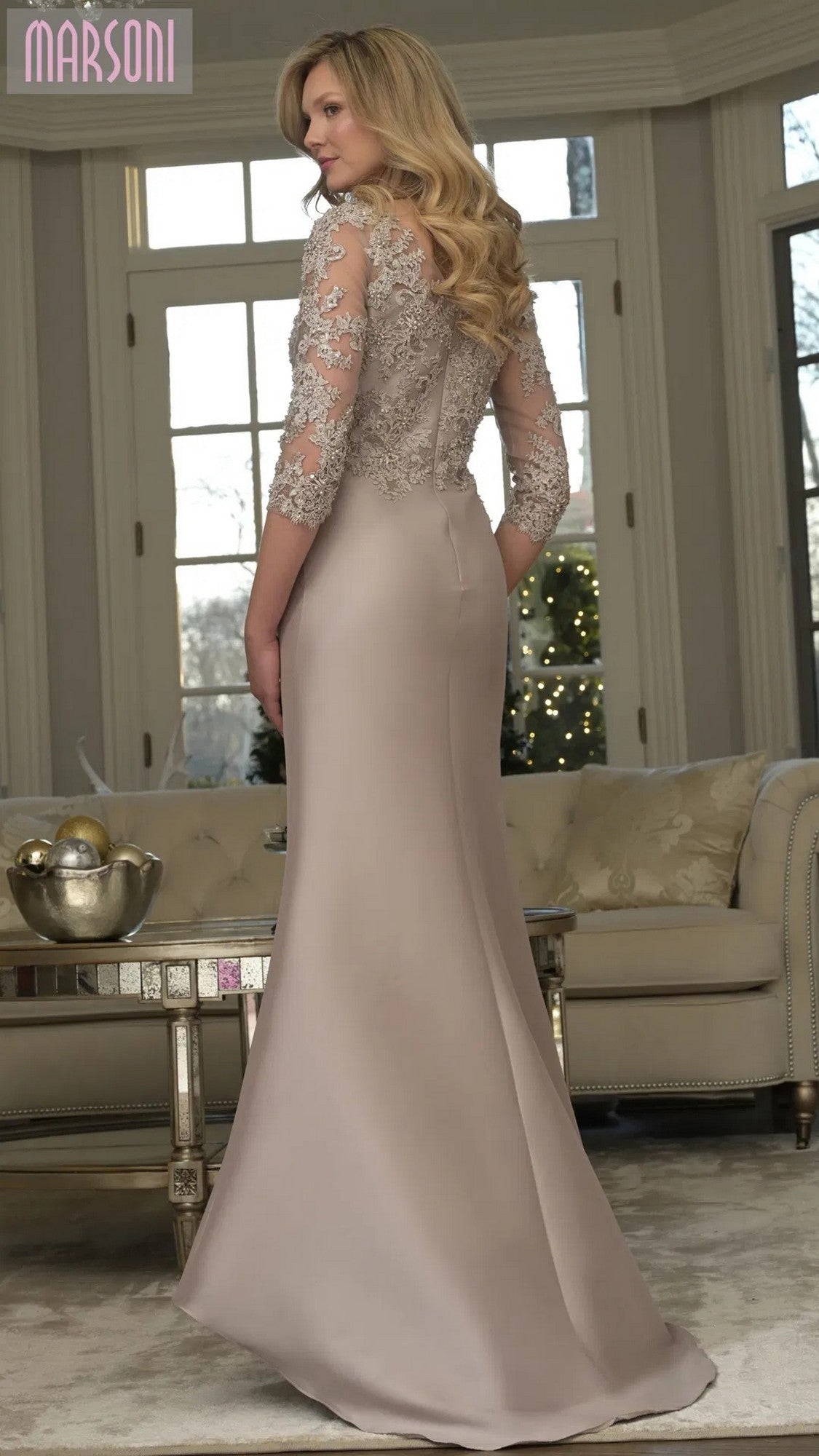 A woman in the Marsoni MV1357 long formal dress with lace sleeves stands in an elegant living room featuring large windows and neutral decor.